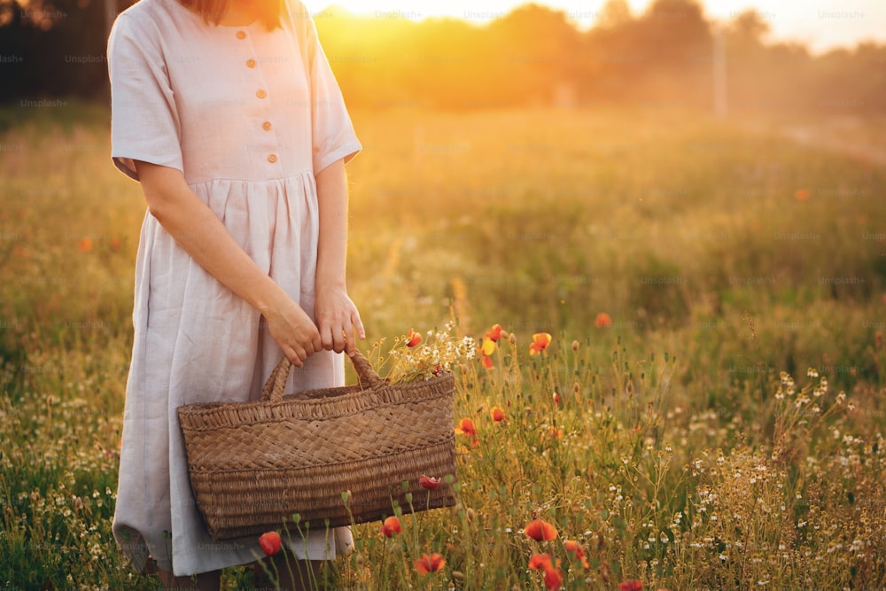 Stylish girl in linen dress holding rustic straw basket with poppy flowers in meadow in sunset light. Boho woman relaxing and gathering wildflowers in summer field. Atmospheric moment