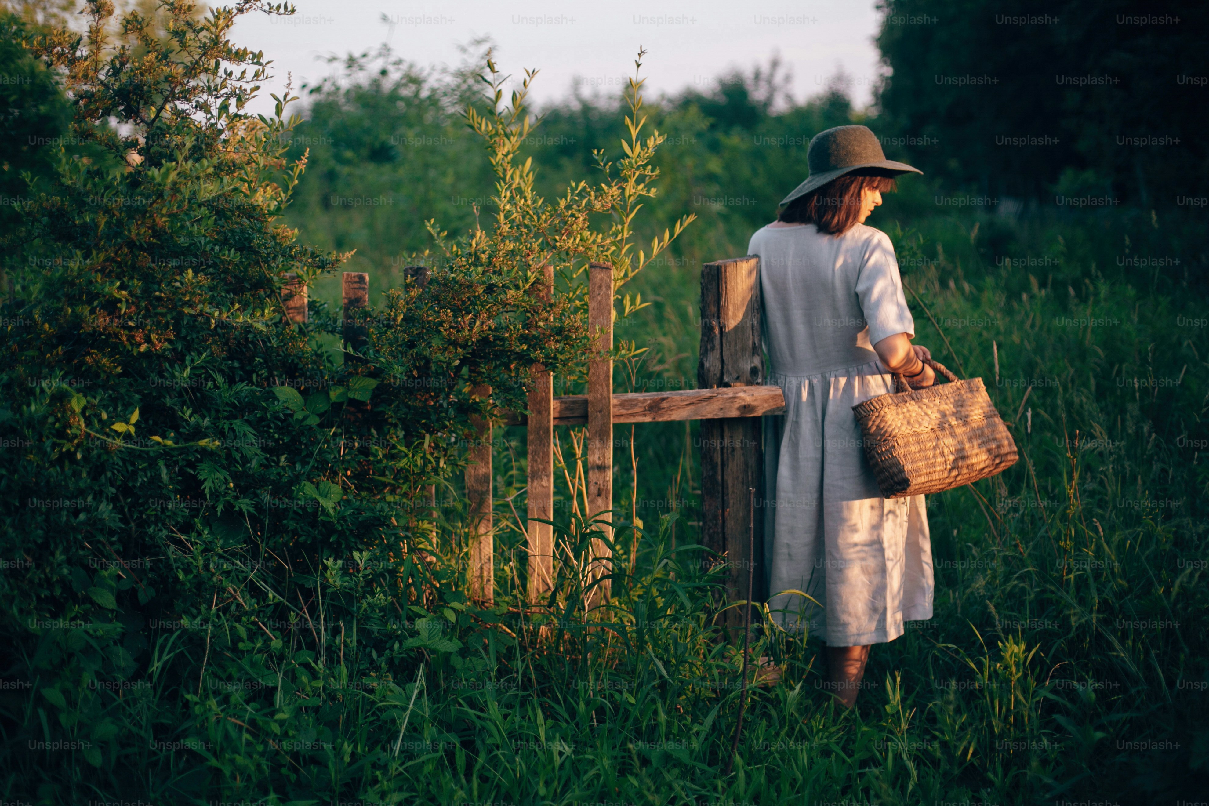 Stylish girl in linen dress holding rustic straw basket at wooden fence  in sunset light. Boho woman relaxing and gathering wildflowers in summer countryside. Atmospheric rural moment