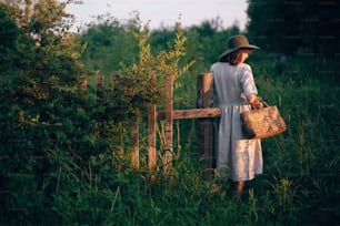 Stylish girl in linen dress holding rustic straw basket at wooden fence  in sunset light. Boho woman relaxing and gathering wildflowers in summer countryside. Atmospheric rural moment