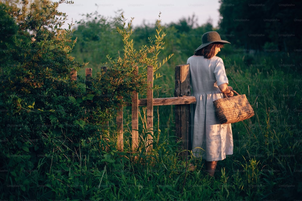 Stilvolles Mädchen im Leinenkleid, das rustikalen Strohkorb am Holzzaun im Sonnenuntergangslicht hält. Boho-Frau entspannt sich und sammelt Wildblumen in der Sommerlandschaft. Atmosphärischer ländlicher Moment