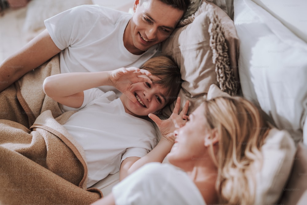 Cheerful kid lying in bed between his parents and having fun while putting hands up pretending being scary