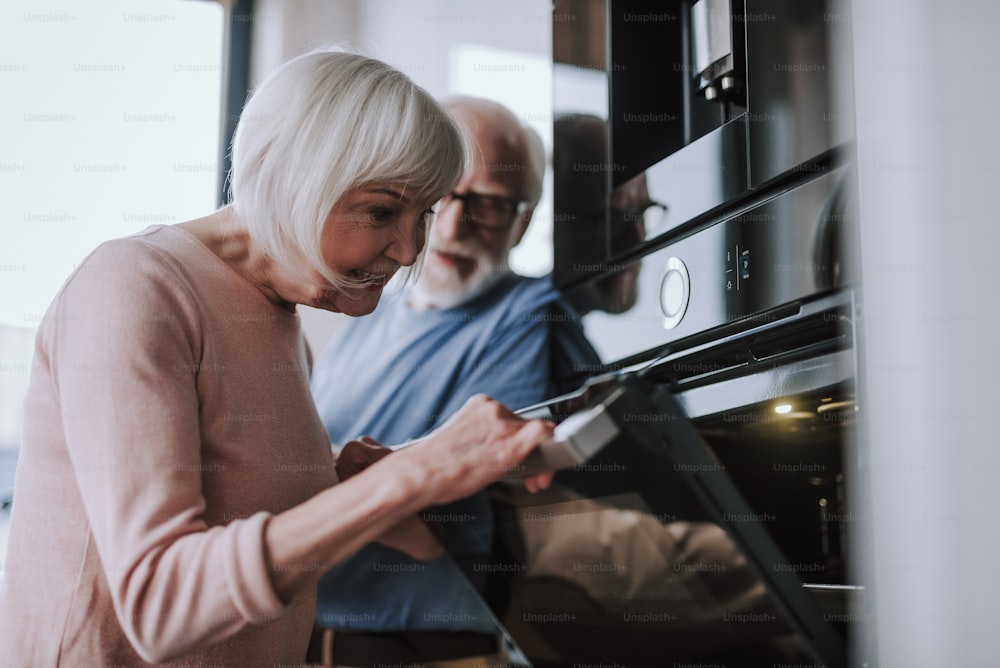 Enjoying time every moment. Waist up low angle portrait of gray haired smiling woman looking into oven to check bakery while her husband ready to help her