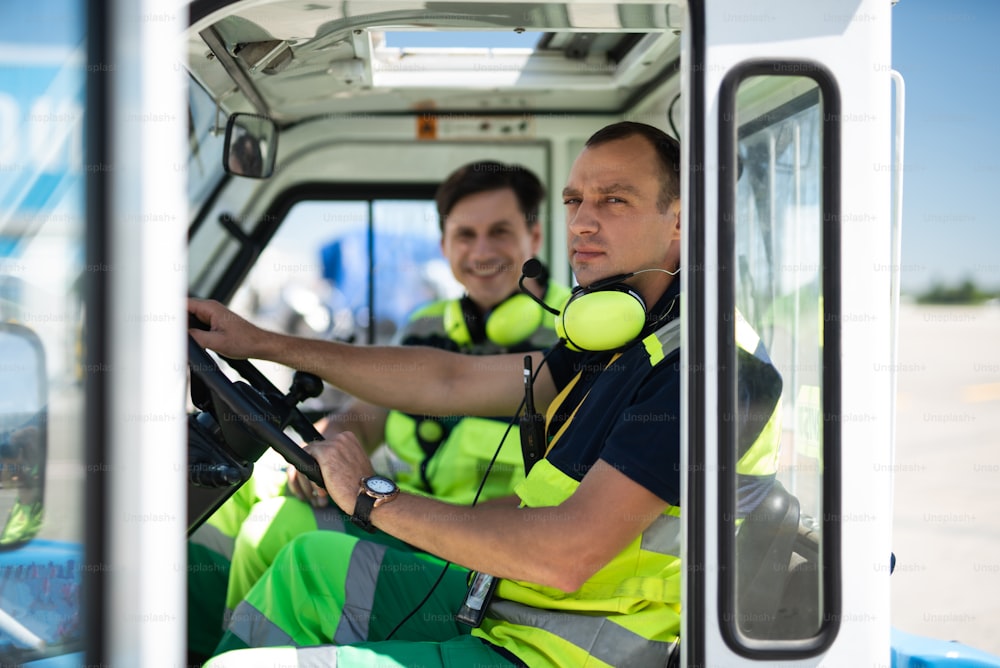 Photoshoot during work. Serious man in headphones with microphone sitting behind the wheel. Smiling colleague on blurred background