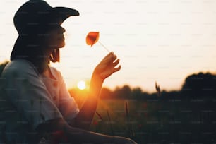 素朴なわらかごに花を咲かせて、太陽の光の中で牧草地にケシの花を咲かせているリネンのドレスを着たスタイリッシュな女の子のシルエット。夏の夕焼け野原でリラックスする帽子をかぶった自由奔放に生きる女性。大気