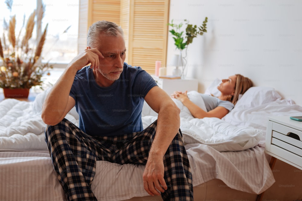 Near sleeping wife. Grey-haired husband wearing pajamas feeling thoughtful sitting near sleeping wife