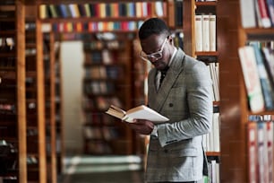 Smart looking african american businessman in glasses standing and reading a book in library.
