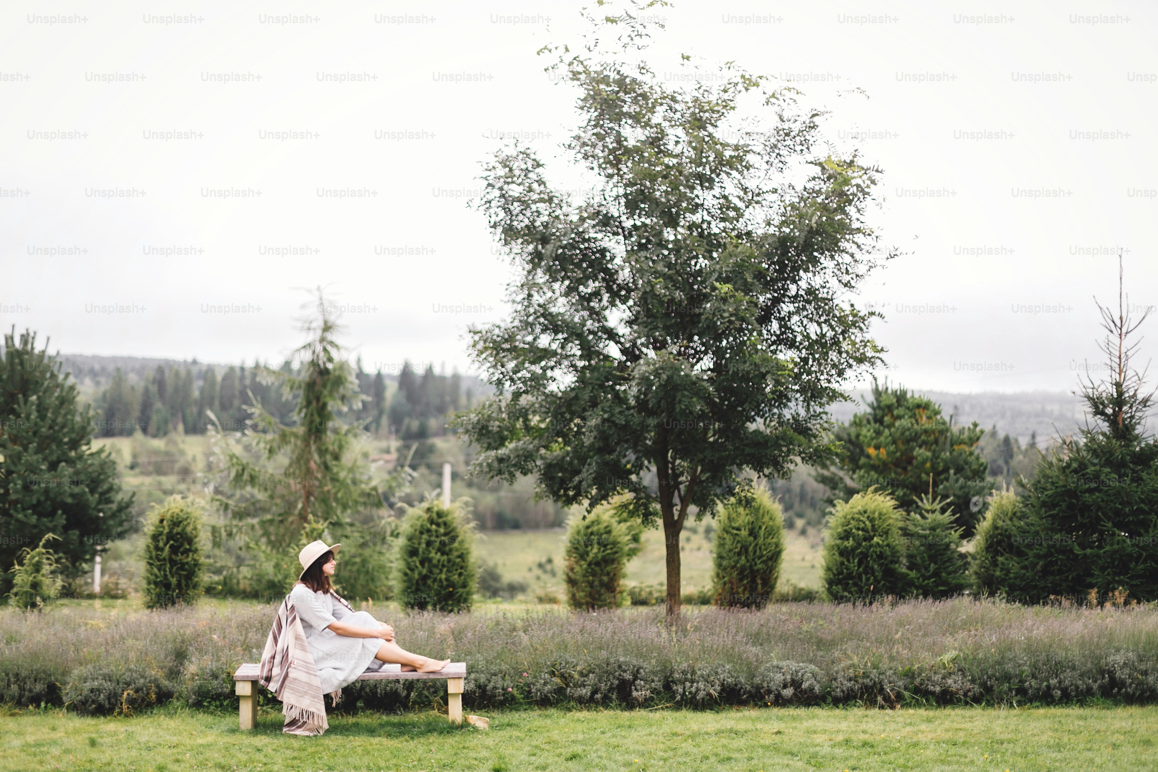 Stylish hipster girl in linen dress and hat sitting on bench at lavender field and relaxing in the morning. Happy bohemian woman enjoying vacation in mountains. Atmospheric rustic moment