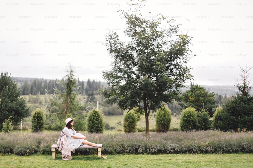 Stylish hipster girl in linen dress and hat sitting on bench at lavender field and relaxing in the morning. Happy bohemian woman enjoying vacation in mountains. Atmospheric rustic moment