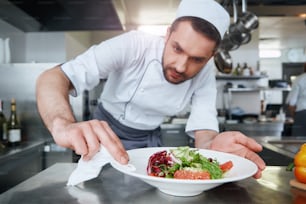 Dark-haired caucasian chef wearing white uniform finishes cooking salad. He wipes the sides of the plate to make dish look better