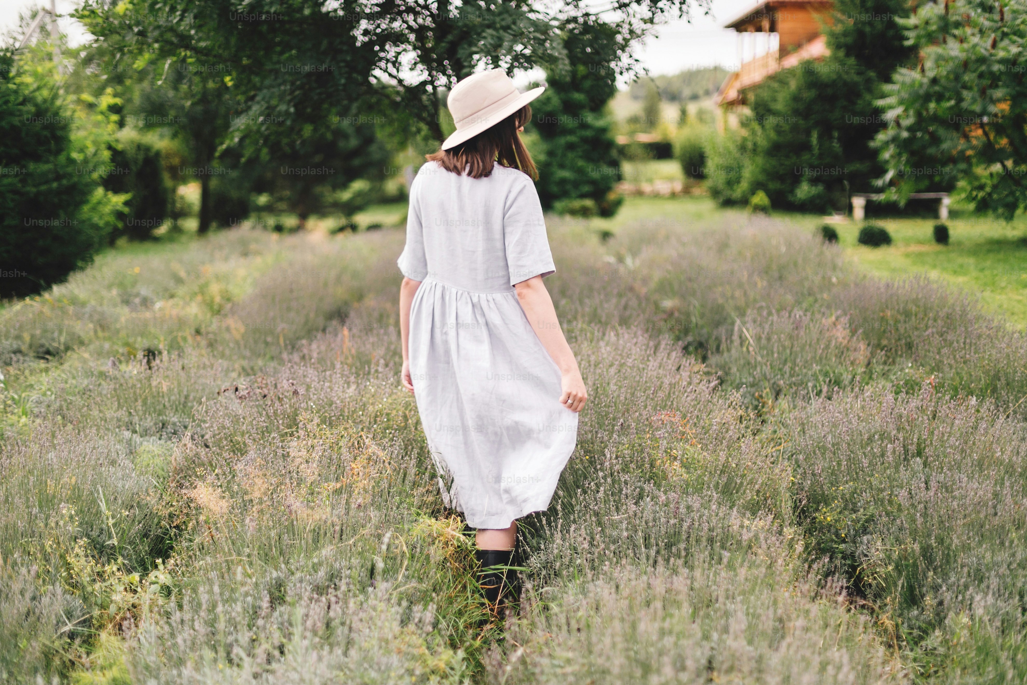 Stylish hipster girl in linen dress and hat walking in lavender field and relaxing. Happy bohemian woman enjoying lavender aroma. Back view. Atmospheric calm rural moment. Space for text
