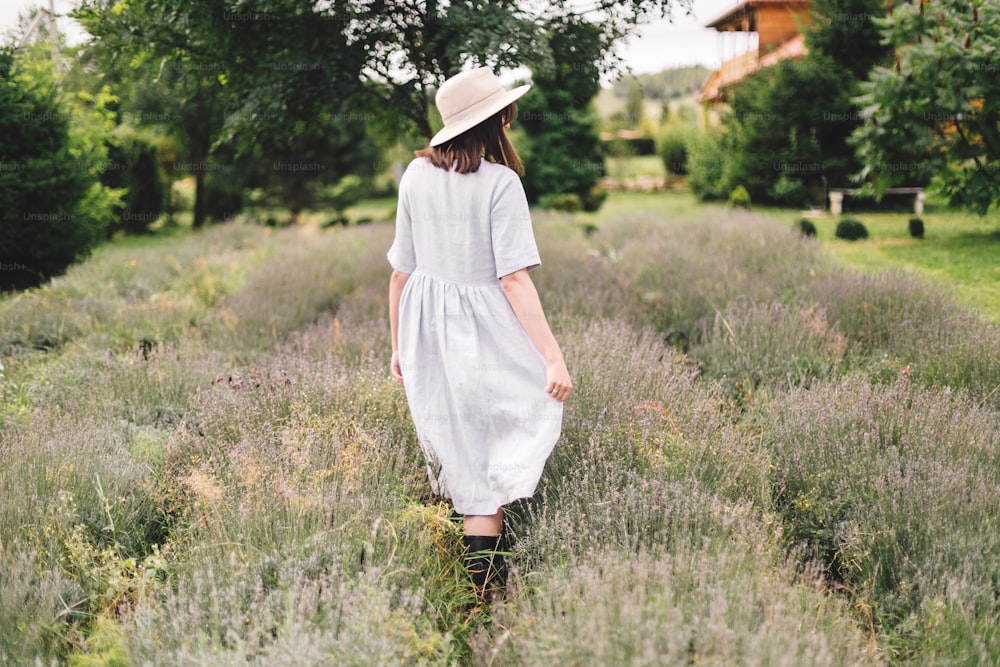 リネンのドレスと帽子をかぶったスタイリッシュなヒップスターの女の子がラベンダー畑を歩き、リラックスしています。ラベンダーの香りを楽しむ幸せなボヘミアン女性。背面図。雰囲気のある穏やかな田舎の瞬間。テキスト用のスペース