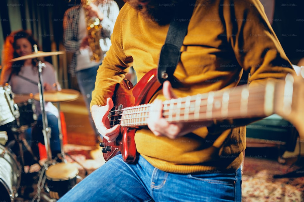 Close up of bass guitarist playing guitar while sitting on the chair. In background saxophonist and piano player. Home studio interior.