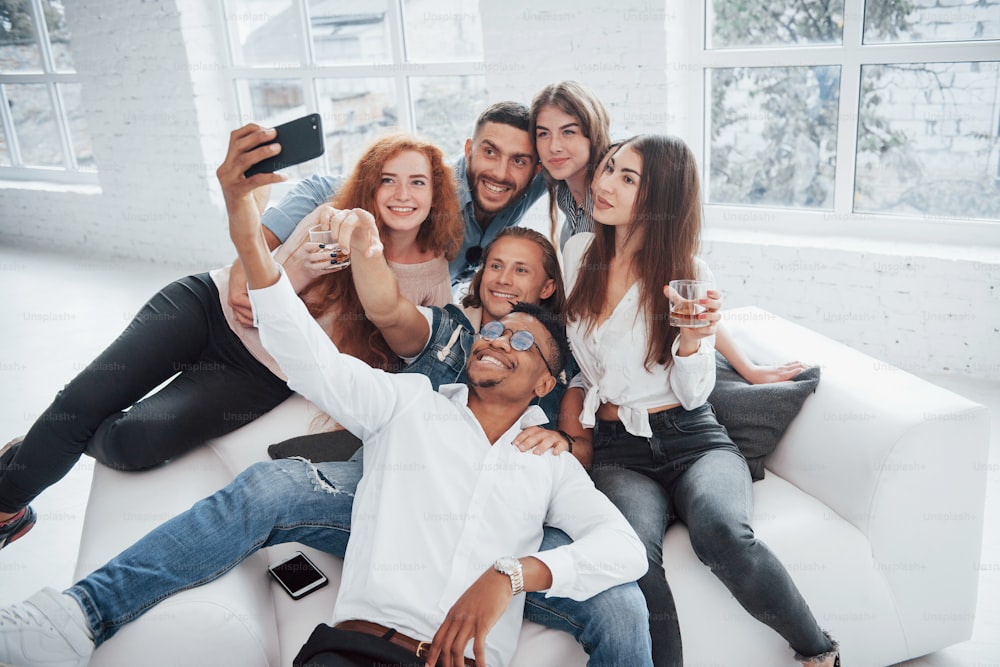 Hacer selfies. Jóvenes amigos alegres divirtiéndose y bebiendo en el interior blanco.