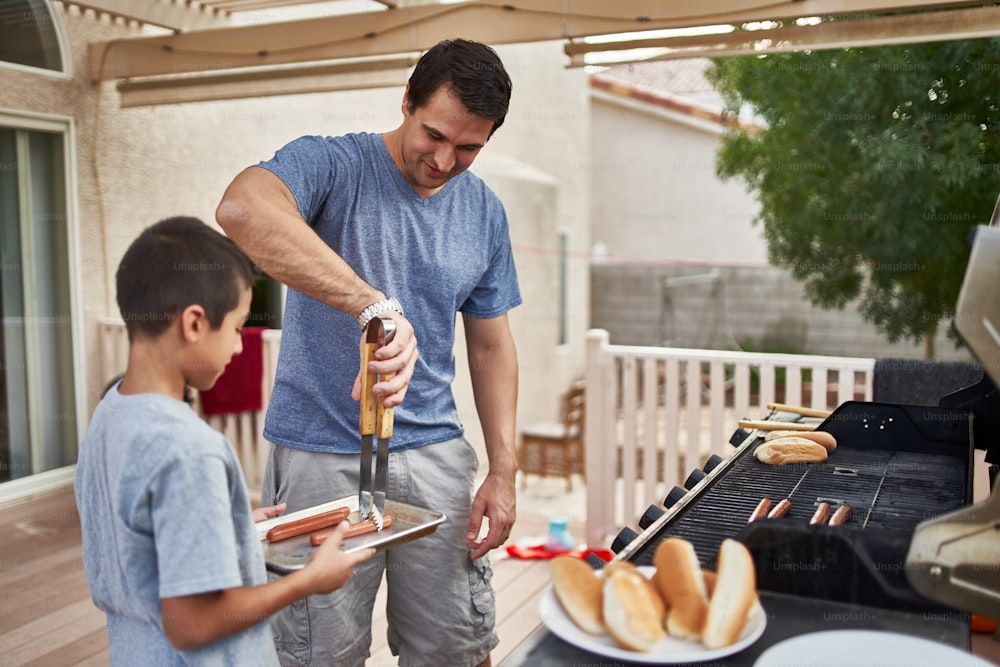 Padre e figlio che grigliano hot dog insieme sulla griglia a gas del cortile durante il giorno