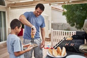 father and son grilling hot dogs together on backyard gas grill during the day