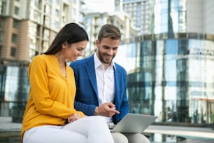 Excited businessman showing presentation on laptop to his colleague. Woman is looking at gadget with interest and smiling with satisfaction. They are sitting outdoor in city