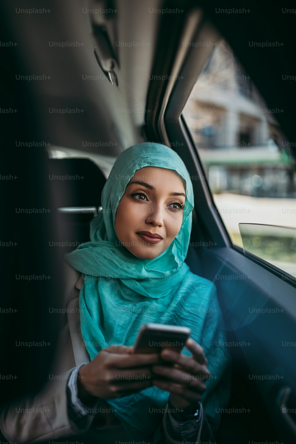 고급 자동차의 뒷좌석에 앉아 히잡을 쓴 아름다운 무슬림 아라비아 여성. 그녀는 스마트 폰을 사용하고 창문을 통해 보았다.