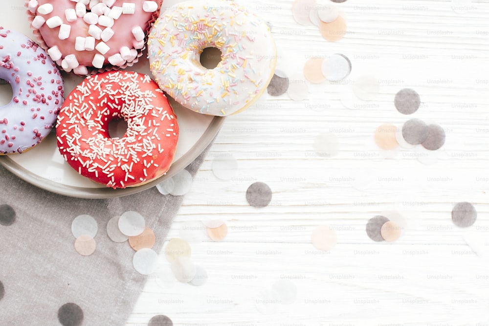 Delicious colorful donuts with sprinkles on stylish plate on white table with confetti, flat lay. Party concept. No diet. Candy bar at wedding reception. Purple and pink donuts