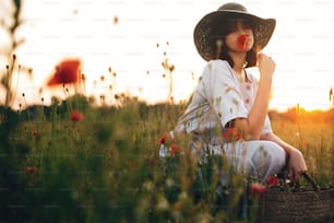 Stylish girl in linen dress smelling poppy flower in meadow in sunset light with flowers in rustic straw basket. Boho woman in hat relaxing in summer field. Atmospheric moment. Space text