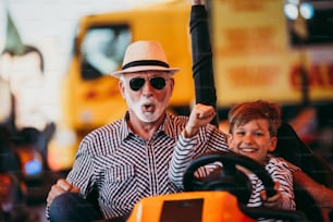 Grandfather and grandson having fun and spending good quality time together in amusement park. They enjoying and smiling while driving bumper car together.