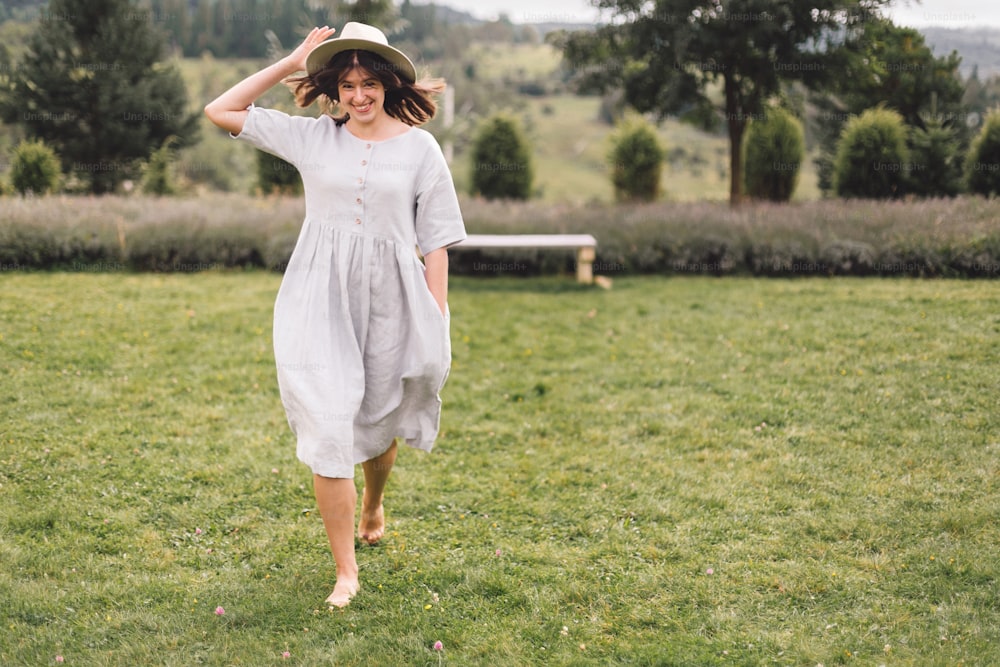 린넨 드레스와 모자를 쓴 세련된 힙스터 소녀는 라벤더 밭에서 즐거운 시간을 보내고 산에서 휴식을 취합니다. 보헤미안 여자는 웃으며 휴가를 즐기고 있다. 분위기 있는 소박한 순간. 복사 공간