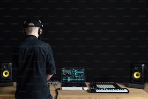 Rear back behind view of unrecognizable man in headset. He making music and using special controller instrument inside house room studio with black dark wall interior with copy empty space for text