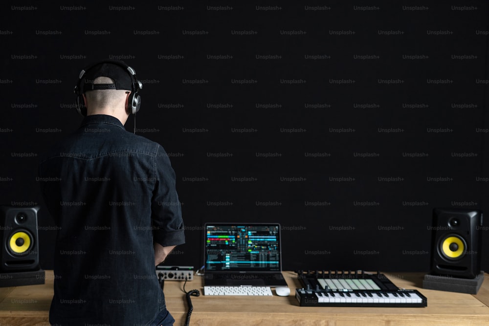 Rear back behind view of unrecognizable man in headset. He making music and using special controller instrument inside house room studio with black dark wall interior with copy empty space for text