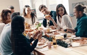 Group of young people in casual wear eating pizza and smiling while having a dinner party indoors