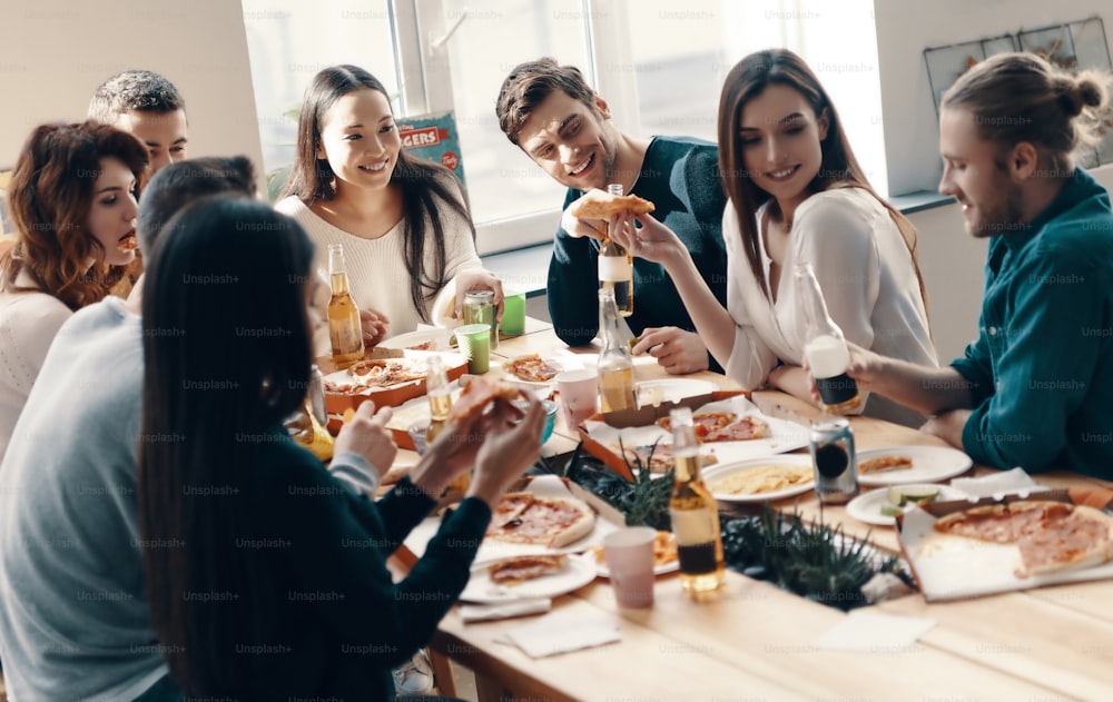 Grupo de jóvenes con ropa informal comiendo pizza y sonriendo mientras tienen una cena en el interior