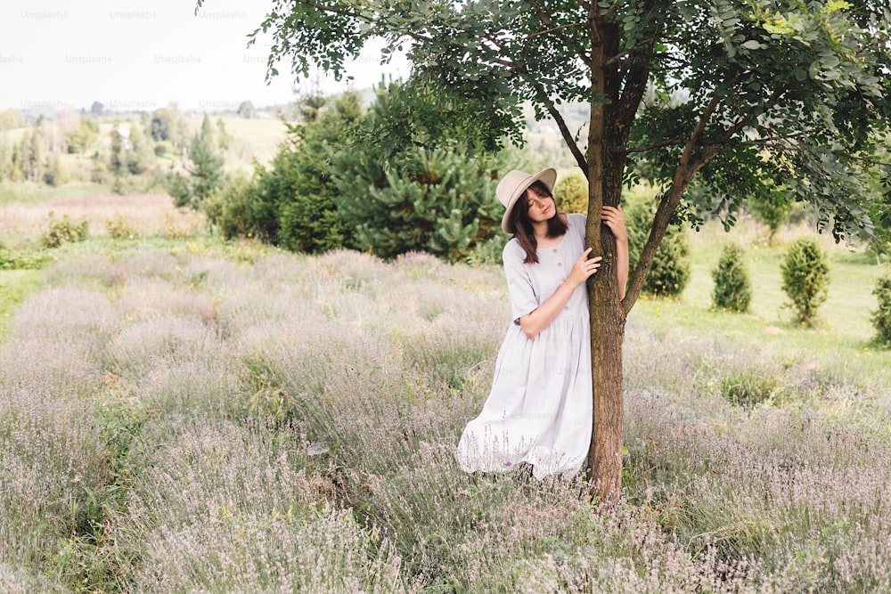 Stylish hipster girl in linen dress and hat relaxing in lavender field near tree. Happy bohemian woman enjoying summer vacation in mountains. Atmospheric calm rural moment. Space for text