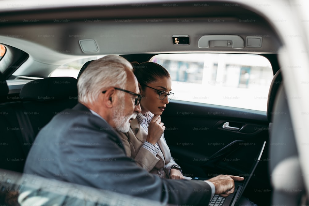 Good looking senior business man and his young woman colleague or coworker sitting on backseat in luxury car. They talking, smiling and using laptop and smart phones. Transportation in corporate business concept.