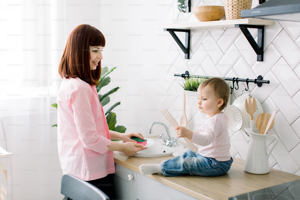 Young happy mother and her cute curly toddler daughter having fun together in a white kitchen. Mother is washing the dishes, plates, while baby girl is sitting near sink and playing with wooden fork