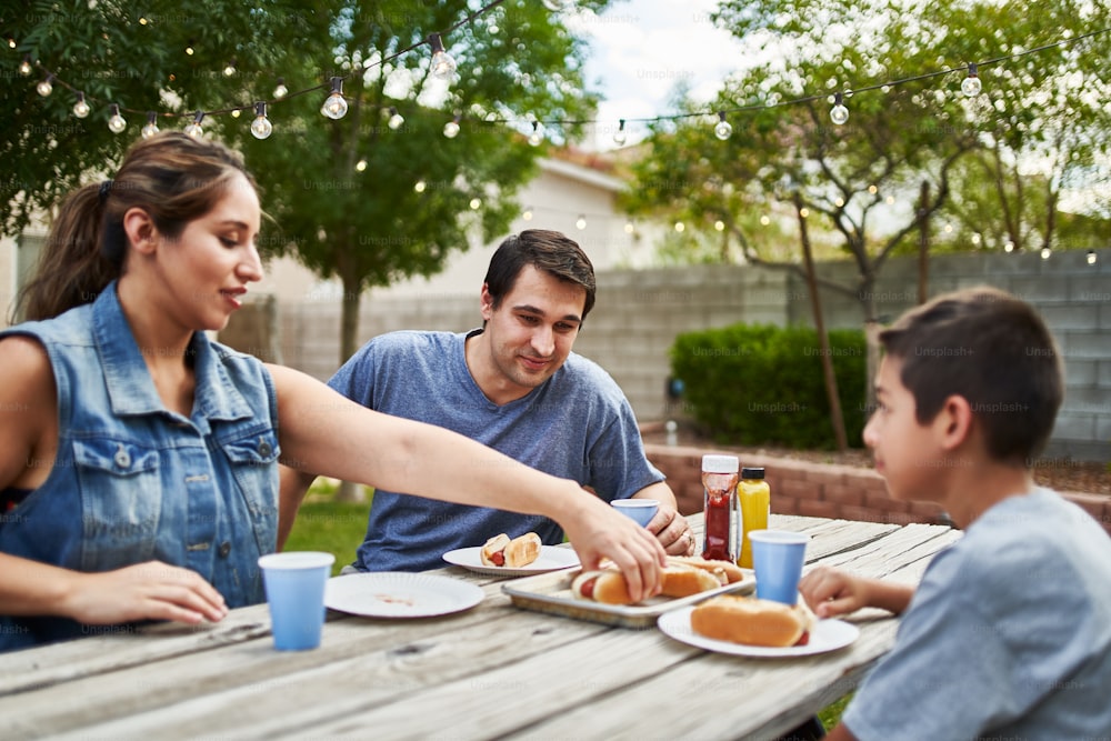 happy hispanic family eating grilled hot dogs on picnic table in backyard during the day