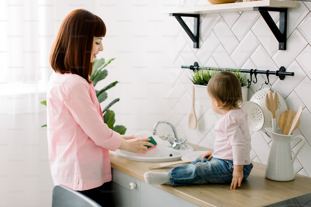 Young happy mother and her cute curly toddler daughter having fun together in a white kitchen. Mother is washing the dishes, plates, while baby girl is sitting near sink and playing with wooden fork