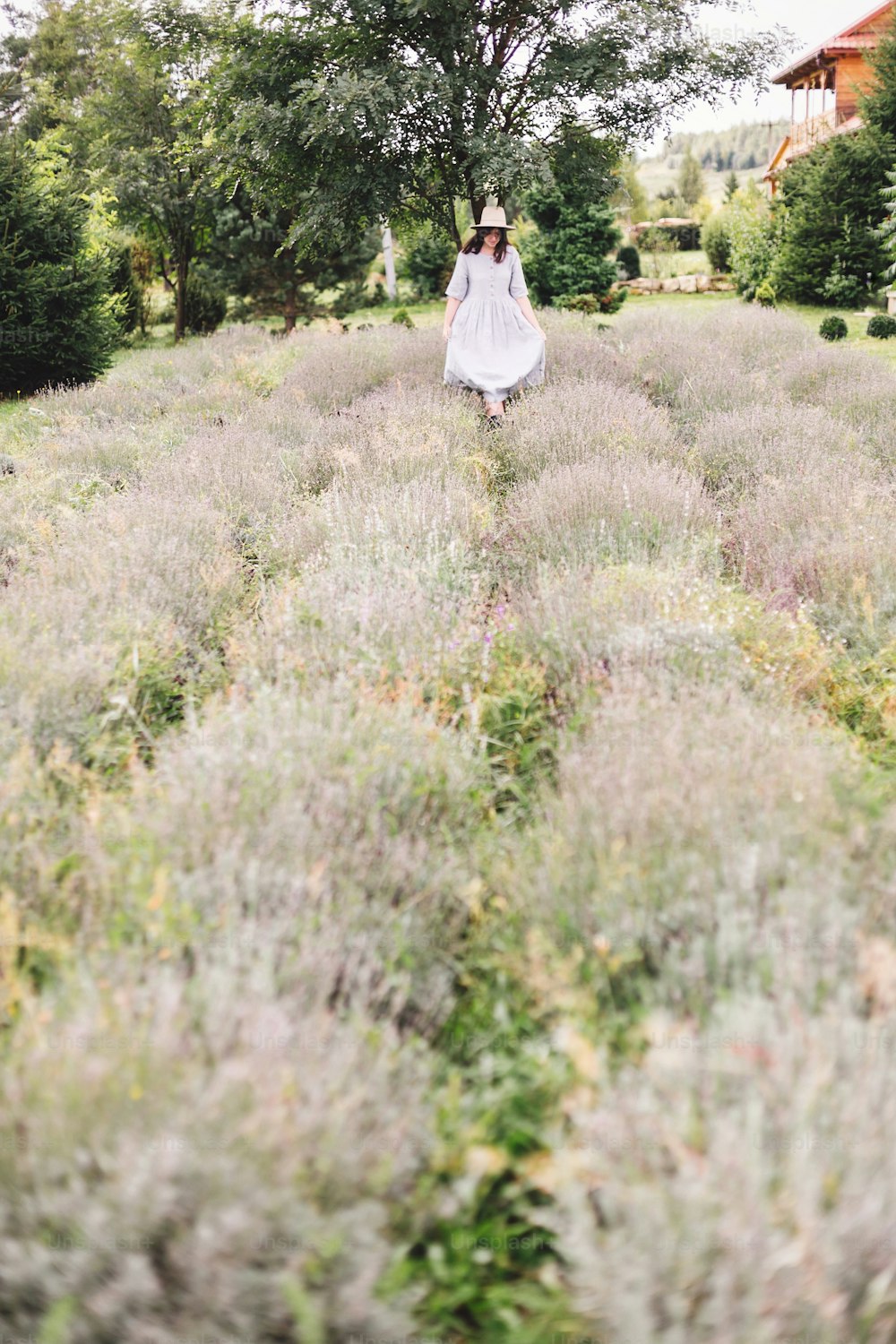 Stylish hipster girl in linen dress and hat walking in lavender field and relaxing. Happy bohemian woman enjoying lavender aroma in summer mountains. Atmospheric calm rural moment