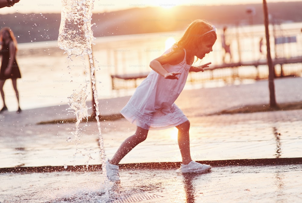 Running through the water. Young girl play in the fountain at the summer heat and lake and woods background.