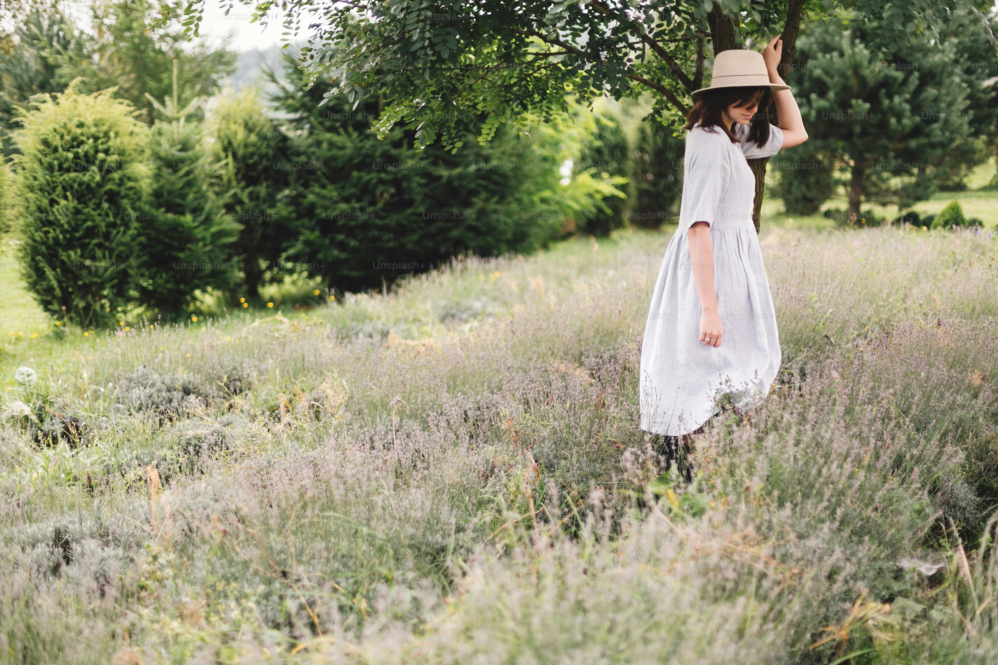 Stylish hipster girl in linen dress and hat relaxing in lavender field near tree. Happy bohemian woman enjoying summer vacation in mountains. Atmospheric calm rural moment. Space for text