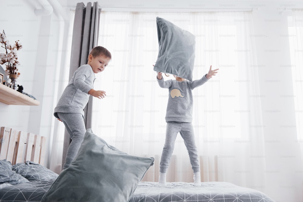 Niños felices jugando en el dormitorio blanco. El niño y la niña, el hermano y la hermana juegan en la cama en pijama. Interior de guardería para niños. Ropa de dormir y ropa de cama para bebés y niños pequeños. Familia en casa