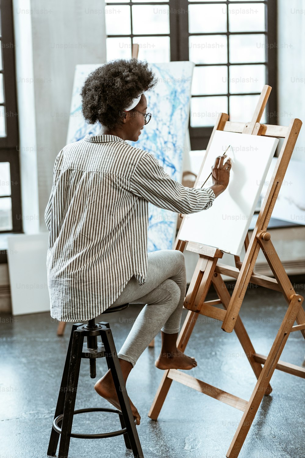 Curly artist. Young curly artist wearing striped shirt sitting on chair and painting on white canvas