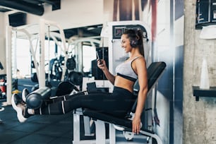 Jeune femme en forme et attrayante qui s’entraîne dans une salle de sport moderne et écoute de la musique avec des écouteurs Bluetooth et un téléphone intelligent.
