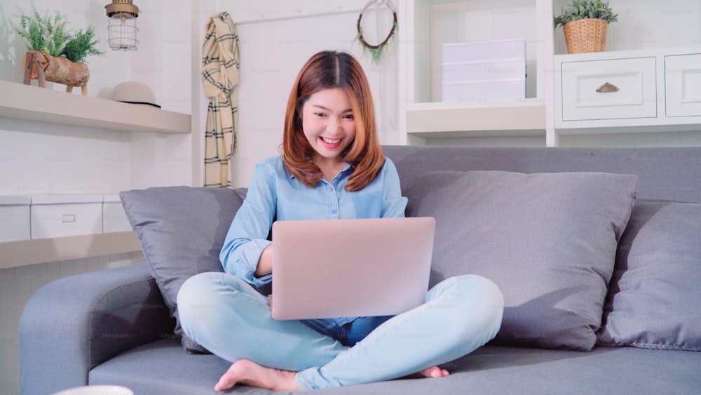 Retrato de una hermosa mujer asiática joven y sonriente atractiva que usa la computadora o la computadora portátil mientras está acostada en el sofá cuando se relaja en la sala de estar de la casa. Disfrutando del tiempo de estilo de vida de las mujeres en el concepto de casa.