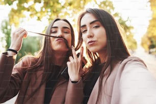Two cheerful and pretty, young brunette twin girls in casual coat having fun while taking selfie and playing with hair, shoving a v sign outdoor in a sunny autumn park on the blurry background.