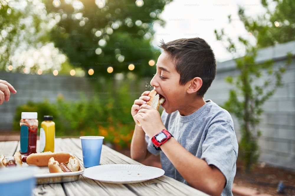 little hispanic boy eating hot dog at family bbq outside in yard during the day