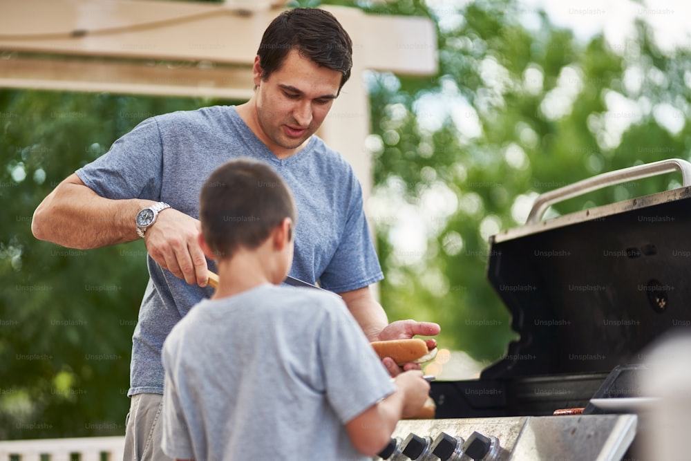 Padre e figlio che grigliano hot dog insieme sulla griglia a gas del cortile durante il giorno