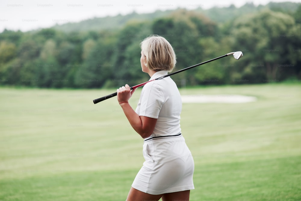 Adult blonde woman have walk on the field while holding the golf stick.