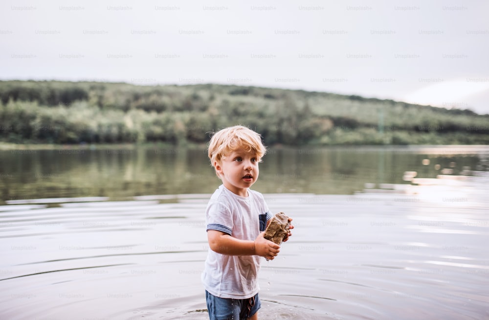 A wet, small toddler boy standing barefoot outdoors in a river in summer, playing with rocks.