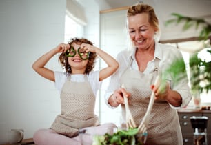 Un retrato de una niña feliz con la abuela preparando ensalada de verduras en casa, divirtiéndose.