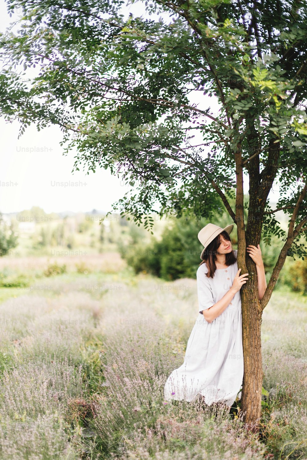 Stylish hipster girl in linen dress and hat relaxing in lavender field near tree. Happy bohemian woman enjoying summer vacation in mountains. Atmospheric calm rural moment. Space for text