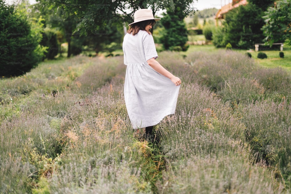 린넨 드레스와 모자를 쓴 세련된 힙스터 소녀가 라벤더 밭을 걷고 휴식을 취하고 있습니다. 여름 산에서 라벤더 향기를 즐기는 행복한 보헤미안 여성. 분위기 있는 차분한 시골의 순간