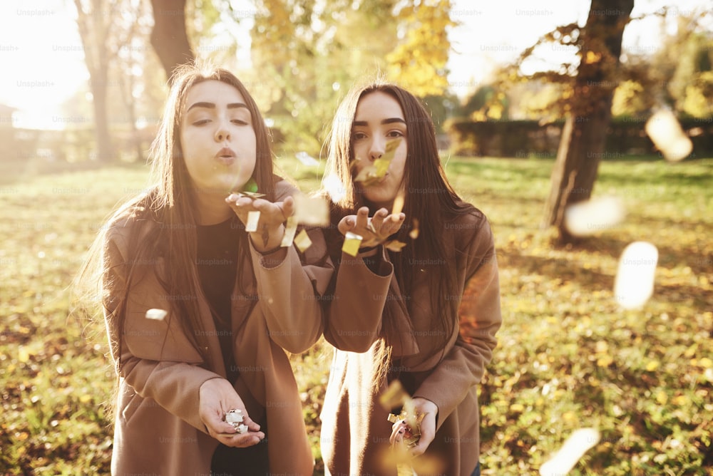 Young brunette twin sisters standing close to each other and blowing confetti into camera, holding some of those in their hands, wearing casual coat in autumn sunny park on blurry background.
